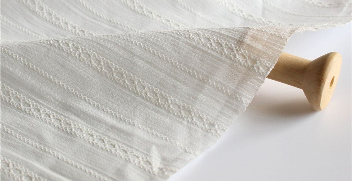 Dimity Cotton Fabric: Soft, Durable, and Timeless Choice A close-up view of a white fabric with delicate stitched patterns, rolled around a wooden spool.