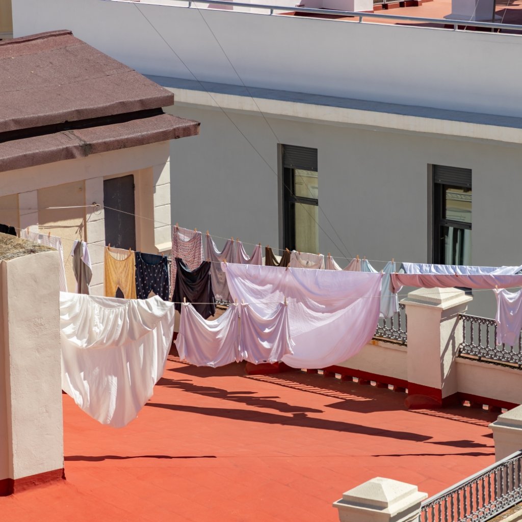 Clotheslines with fresh laundry hanging on a sunlit rooftop terrace, white sheets and colorful garments fluttering against a pale building and red floor.
