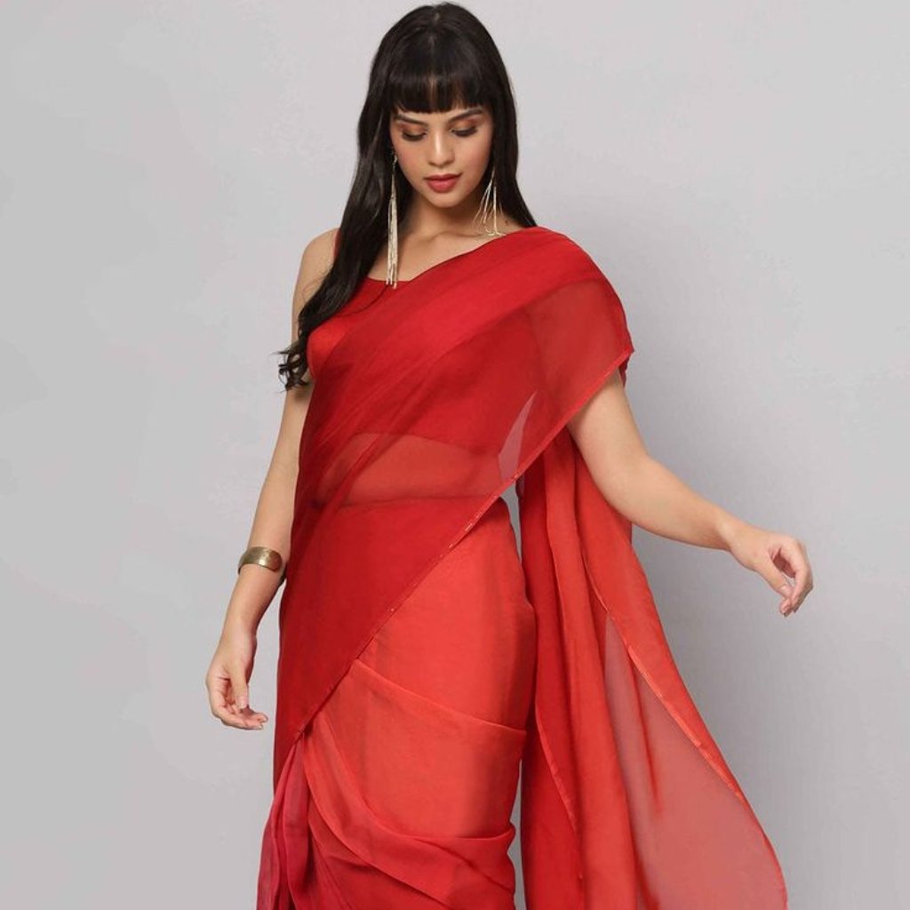 A woman wearing a sheer red saree with an off-shoulder blouse, standing against a light gray background. She has long dark hair with bangs and is accessorized with long earrings and a gold cuff bracelet.