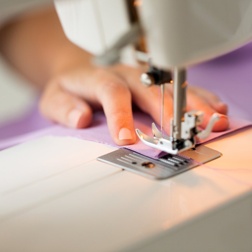 Close-up of a sewing machine stitching a piece of light purple fabric, with a person’s hand guiding the fabric under the presser foot.