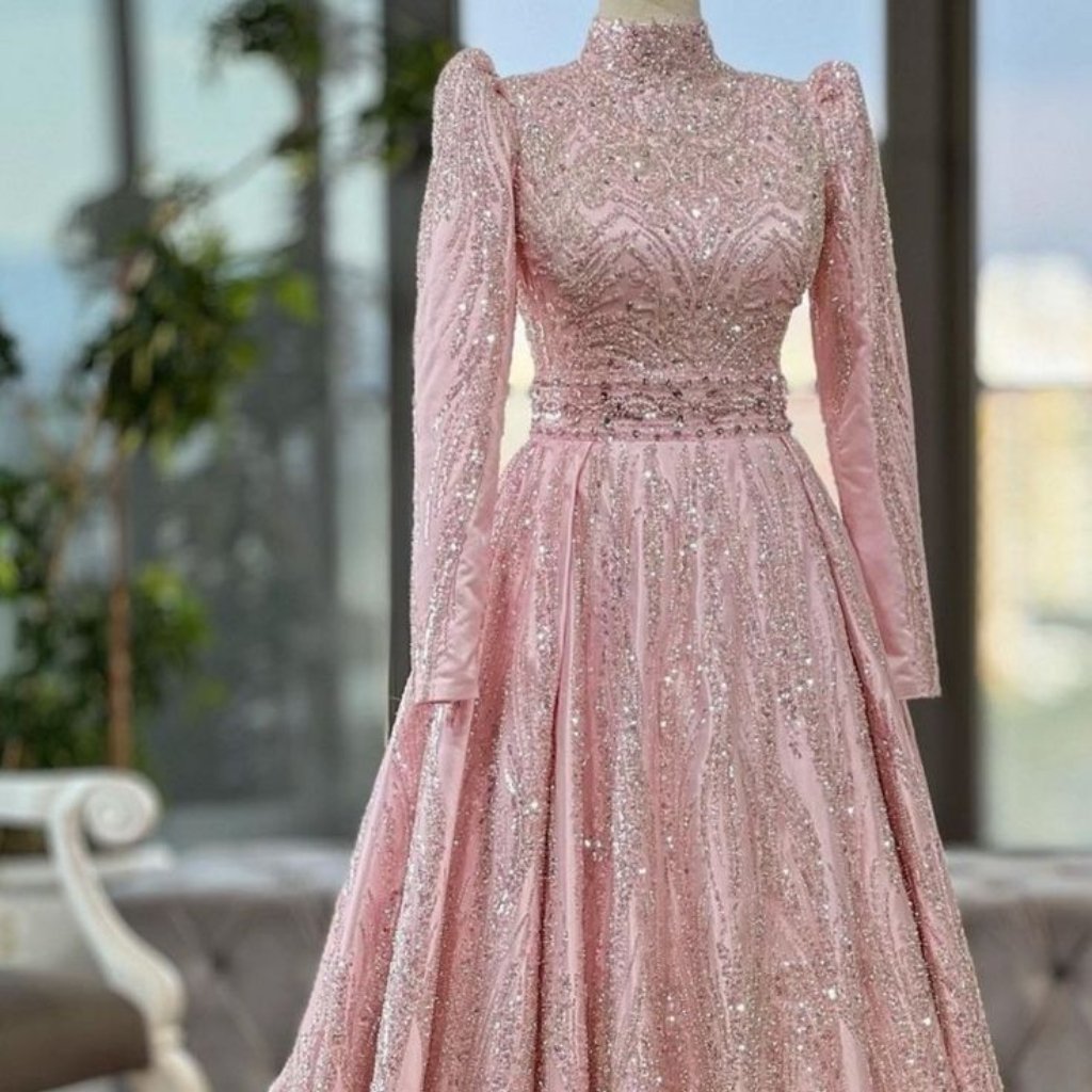 A floor-length, blush-pink evening gown with long sheer sleeves, high lace neckline, and intricate sequined embroidery. The bodice and waist feature ornate beading, and the full, flowing skirt shimmers with glittery details.