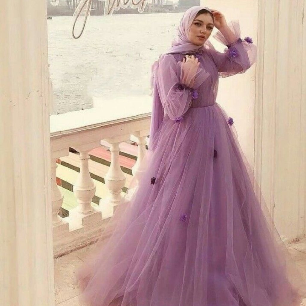 A woman wearing a lilac hijab and a floor-length lavender tulle gown with puff sleeves stands on a balcony by the water.
