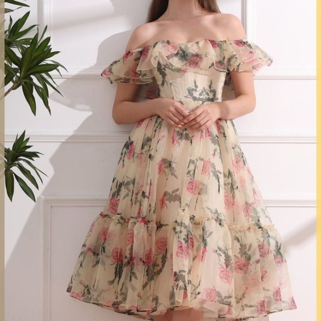 A woman wearing a light, off-the-shoulder floral dress with a tiered, ruffled skirt standing against a white paneled wall.
