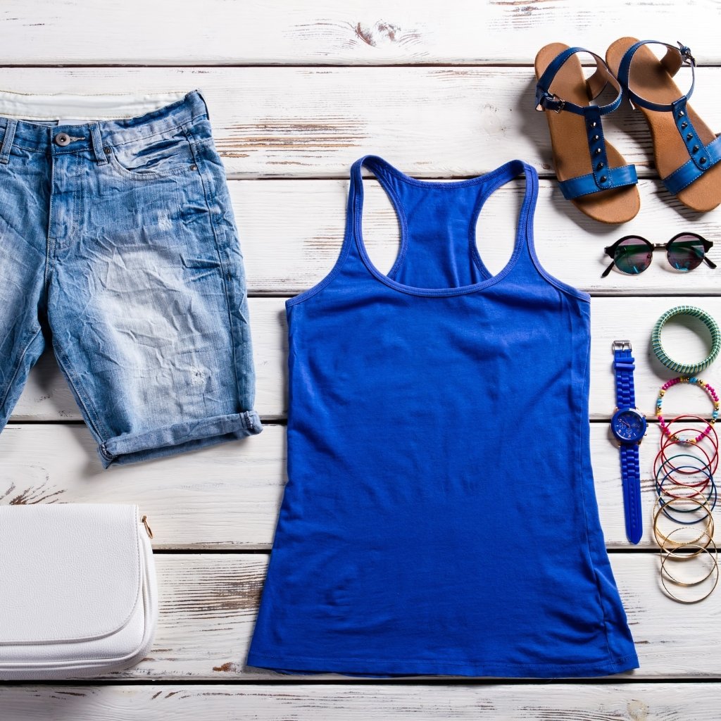 A flat lay of summer clothing and accessories on a white wooden surface: a bright blue tank top, distressed denim shorts, blue sandals, sunglasses, a white purse, and stacked bracelets. The scene emphasizes fabric textures and casual style.