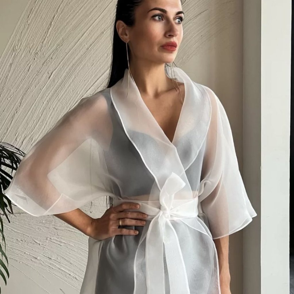 White Organza Fabric: Elegant Style for Dresses and Interiors 29 A woman wearing a sheer white robe with wide, draped sleeves tied at the waist, standing near a light-colored wall.