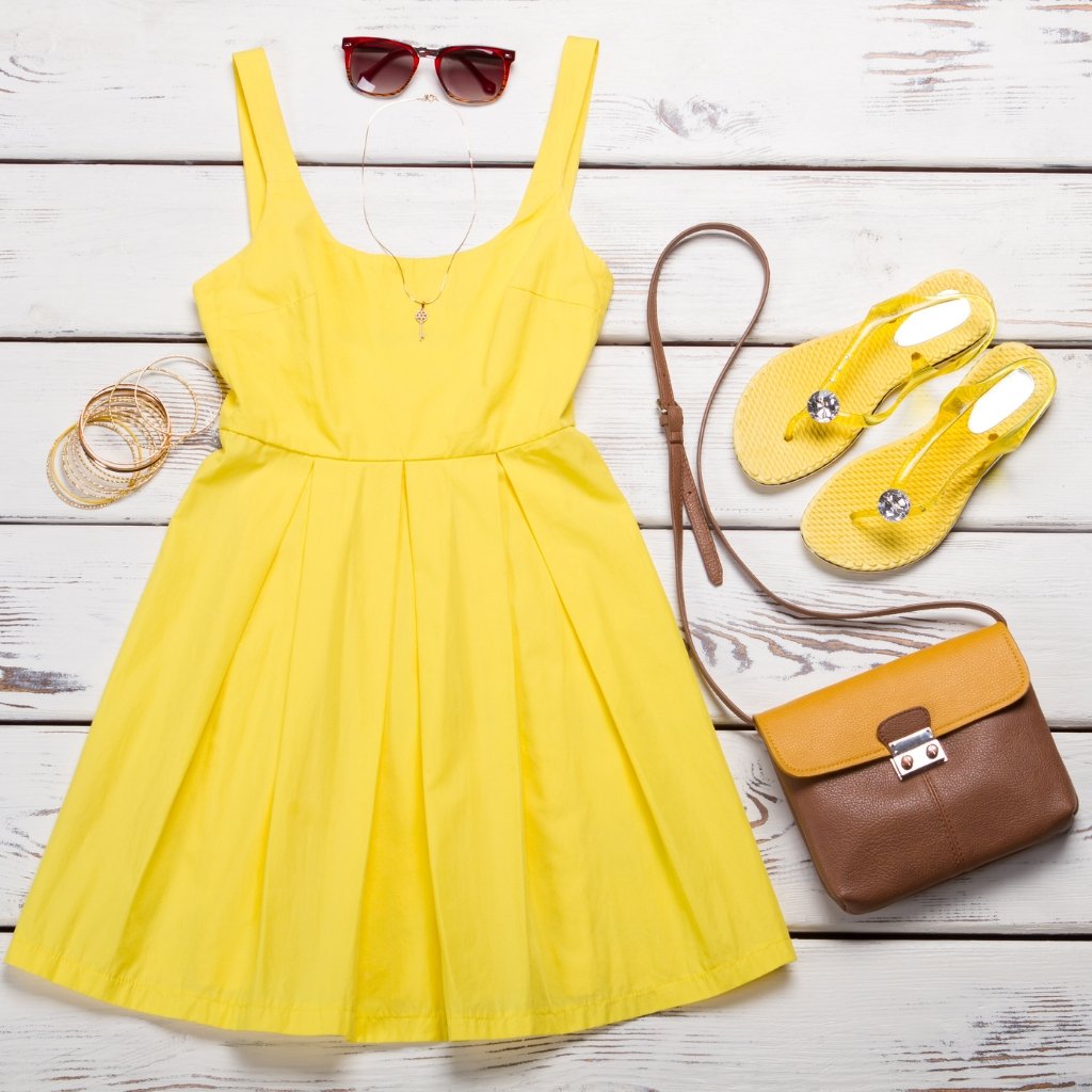 Bright yellow sleeveless dress laid flat on a white wooden surface, with yellow flip-flops, a brown and tan crossbody bag, yellow sunglasses, bracelets, and a pendant necklace arranged around it.