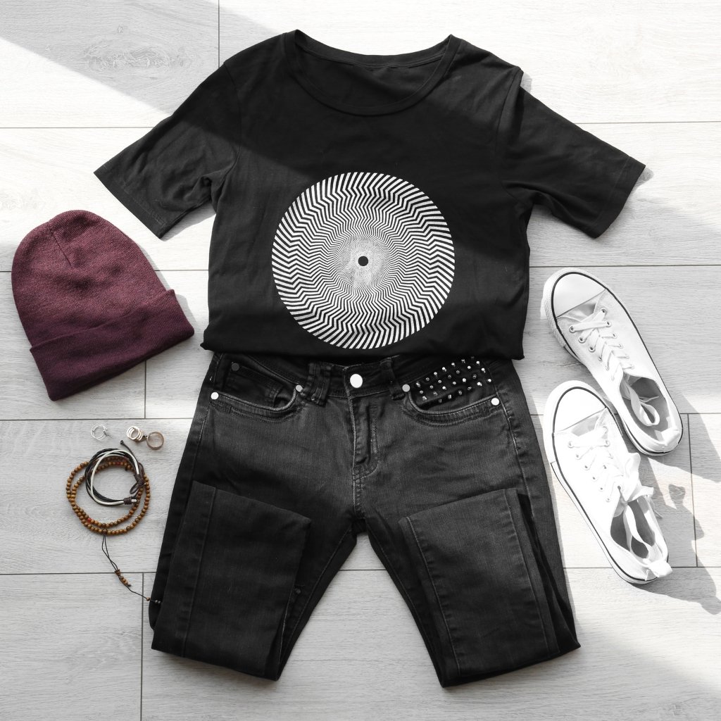 A flat-lay casual outfit on a light floor: black graphic t-shirt with a white concentric circular optical design, paired with black denim shorts featuring studded detailing, white sneakers, a burgundy knit beanie, and several bracelets.