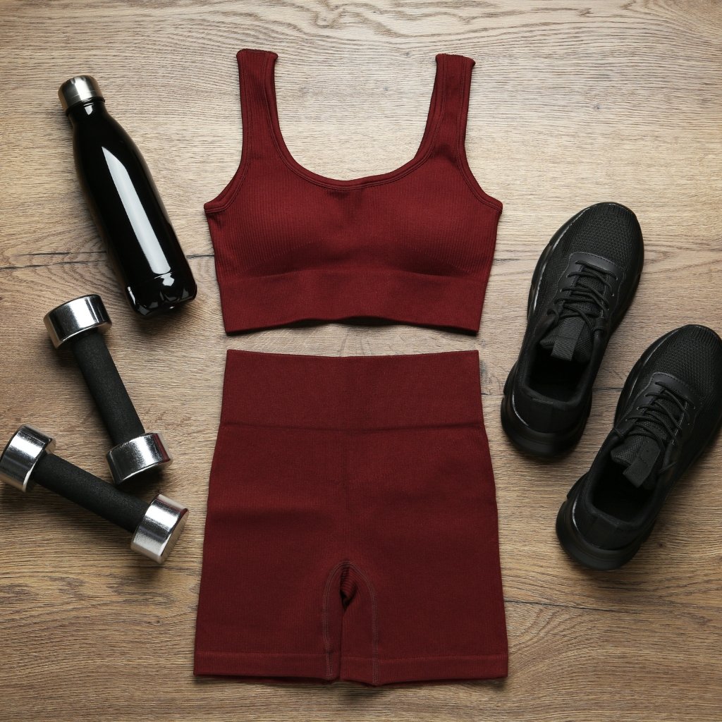 Flat lay of a burgundy sports outfit including a sports bra and high-waisted shorts, black sneakers, a black water bottle, and two dumbbells on a wooden floor.