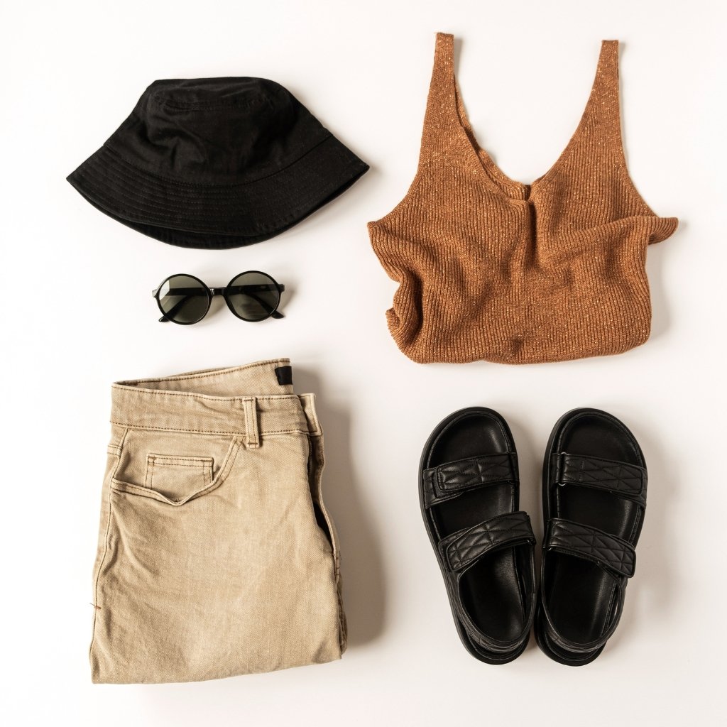 Flat lay of fashion items on a white background: a black bucket hat, round black sunglasses, beige folded shorts, a copper-brown knit tank top, and black sandals arranged neatly.