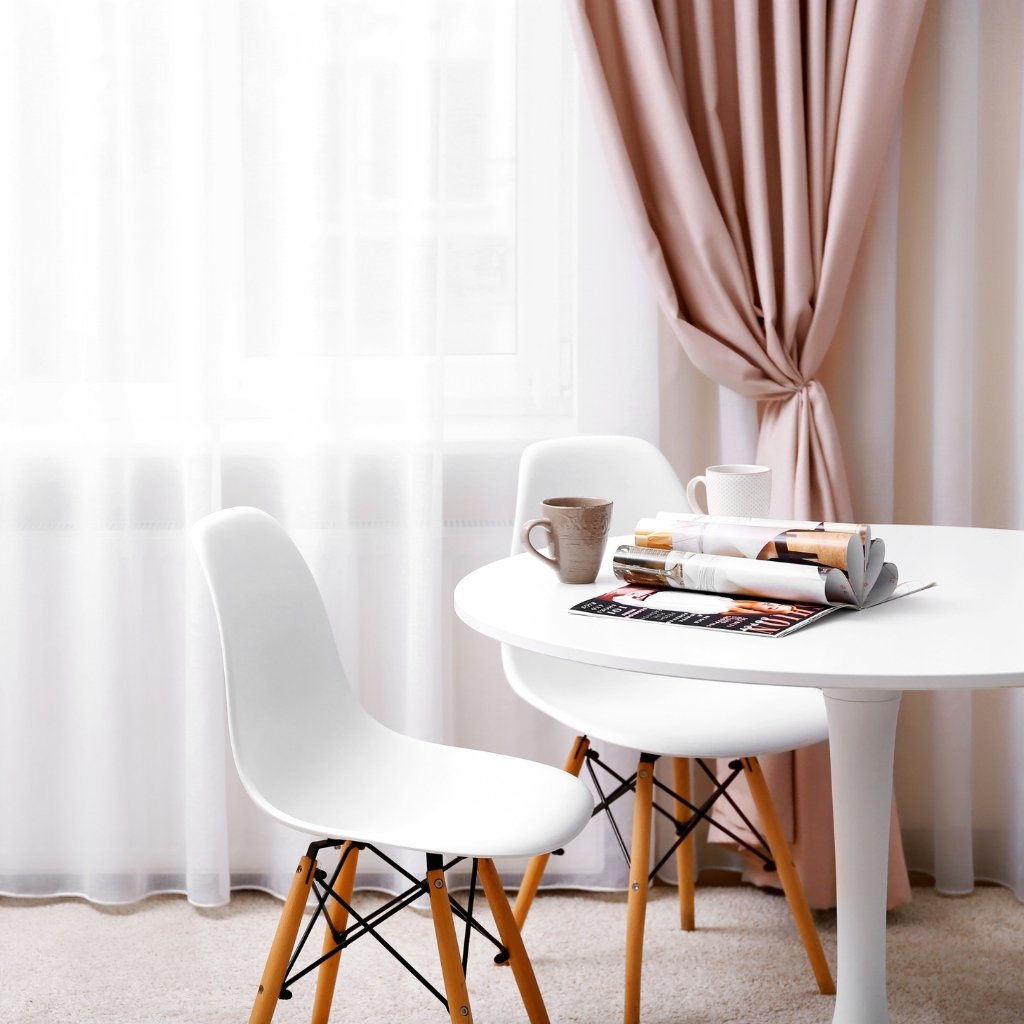 A bright, minimalist dining nook with a white round table, white chairs with wooden legs, an open magazine on the table, and soft pink curtains by a window.