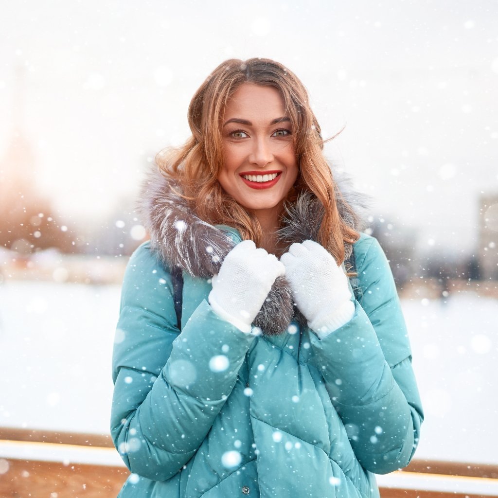 A smiling woman with light brown hair wearing a teal winter coat with a fur-lined hood and white gloves, standing in a snowy outdoor setting with soft falling snow in the foreground and background.
