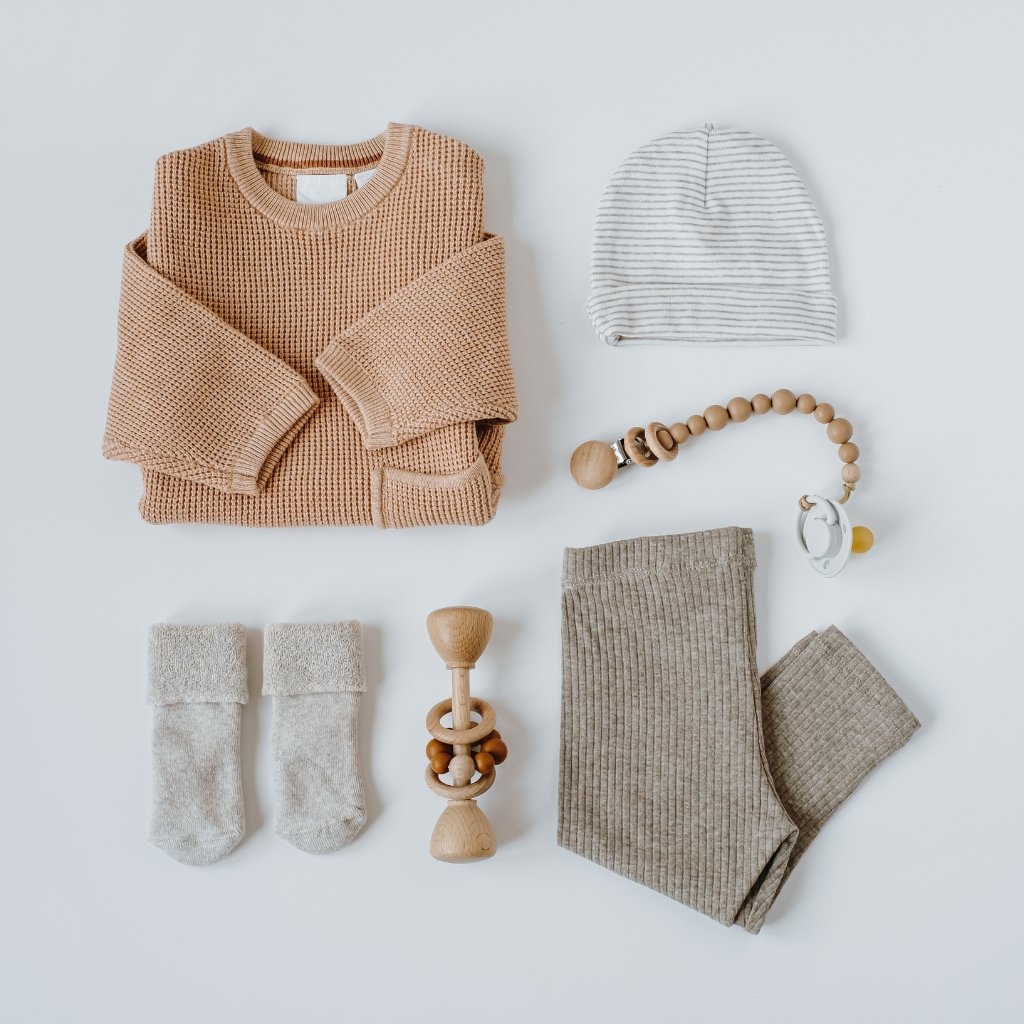 A flat lay of baby essentials: a peach-colored knitted sweater, a striped beanie, two small soft socks, a wooden teething toy, a wooden bead pacifier clip, a pair of corduroy pants, and a small grayish knitted set arranged on a light background.