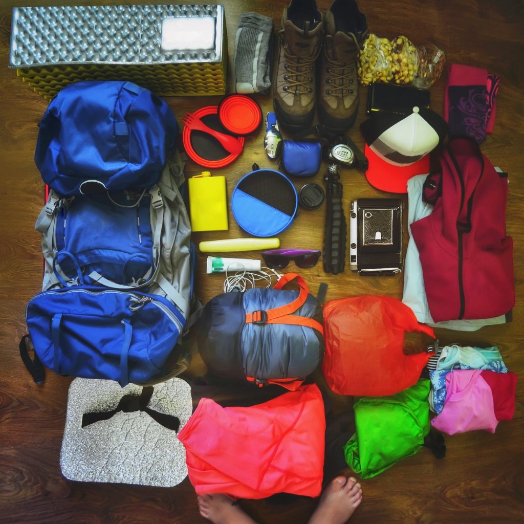 A top-down shot of a well-organized camping gear setup spread on a wooden floor. Items include a blue backpack, hiking boots, a red/pink jacket, a sleeping bag, a foam sleeping pad, a metal box, a yellow water container, a blue stuff sack, red headphones, a cap, a small camera, a blue lunch box, and various colorful bags and clothing. The scene suggests someone packing for a hiking or camping trip.