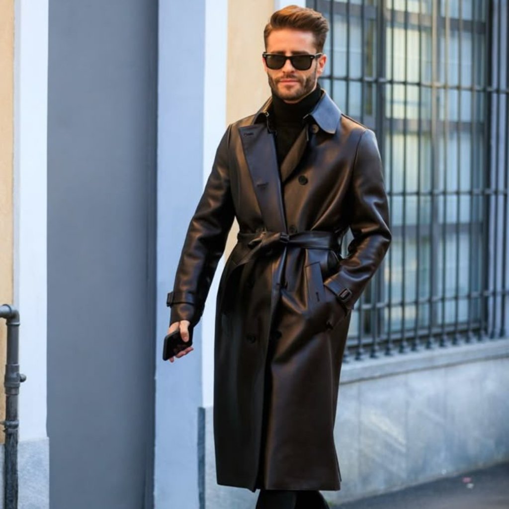 A stylish man wearing a long black leather duster coat, black turtleneck, black pants, sunglasses, and holding a phone as he walks outdoors.
