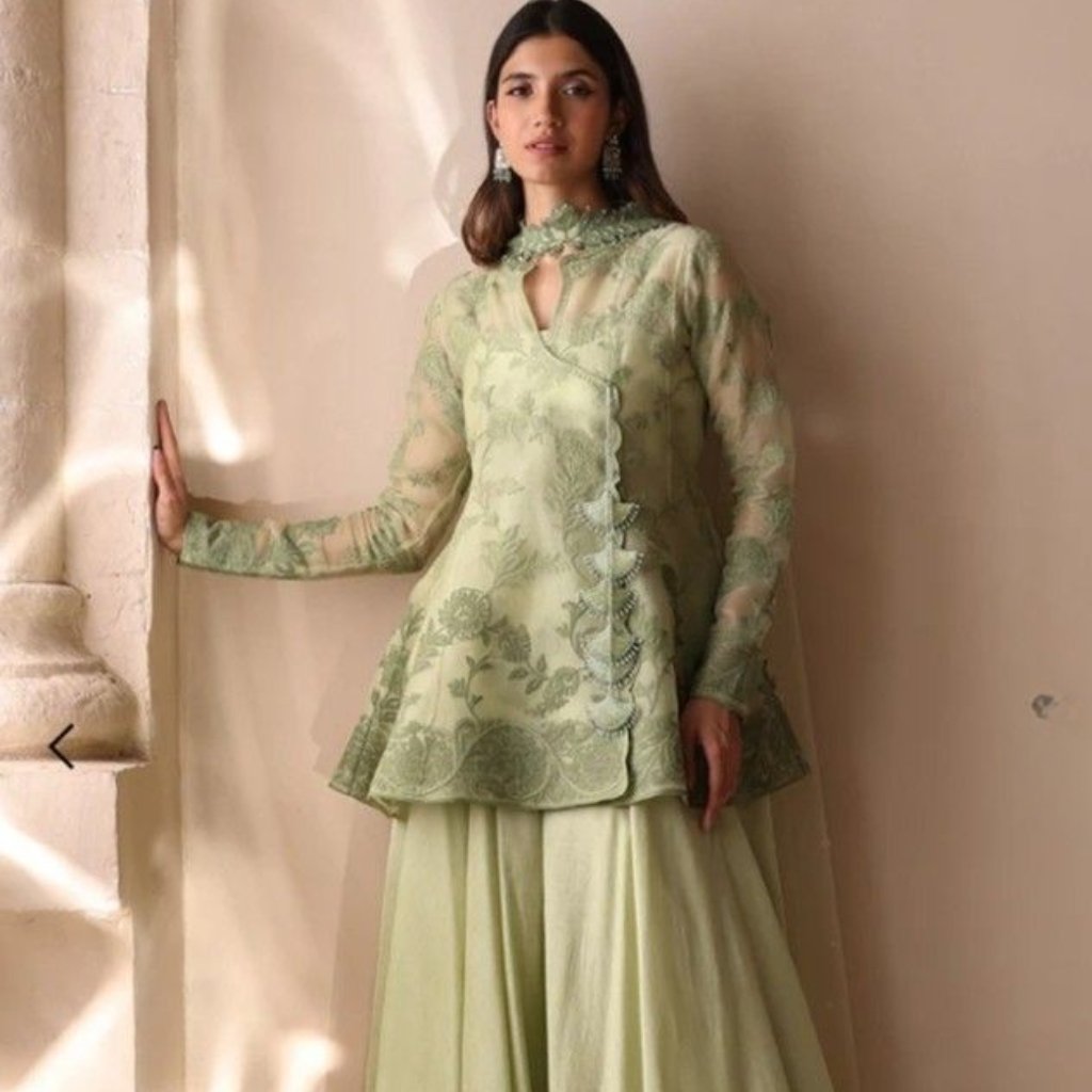 Green Organza Fabric: Elegant Choices for Fashion and Decor 17 A woman stands indoors beside a column, wearing a light sage-green lace top with long sleeves and matching flowing skirt. She gazes slightly to the side, and her hair is loose with statement earrings.