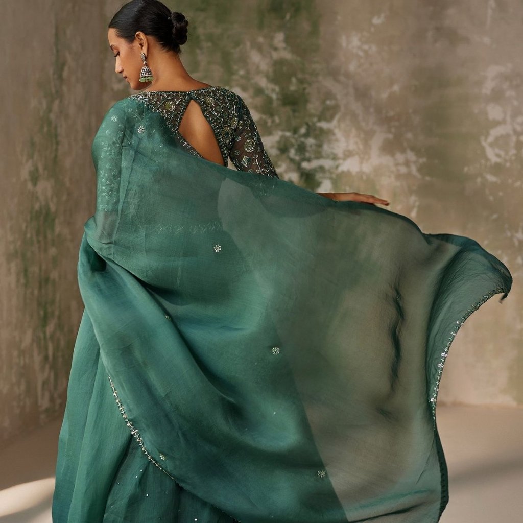 Green Organza Fabric: Elegant Choices for Fashion and Decor 21 A woman wearing a teal-sherwani style outfit, captured from behind as she twirls, her dupatta fluttering gracefully in the air. The back of her embellished blouse features a teardrop cutout, and she accessorizes with large, dangling earrings. The background is a muted, weathered wall.