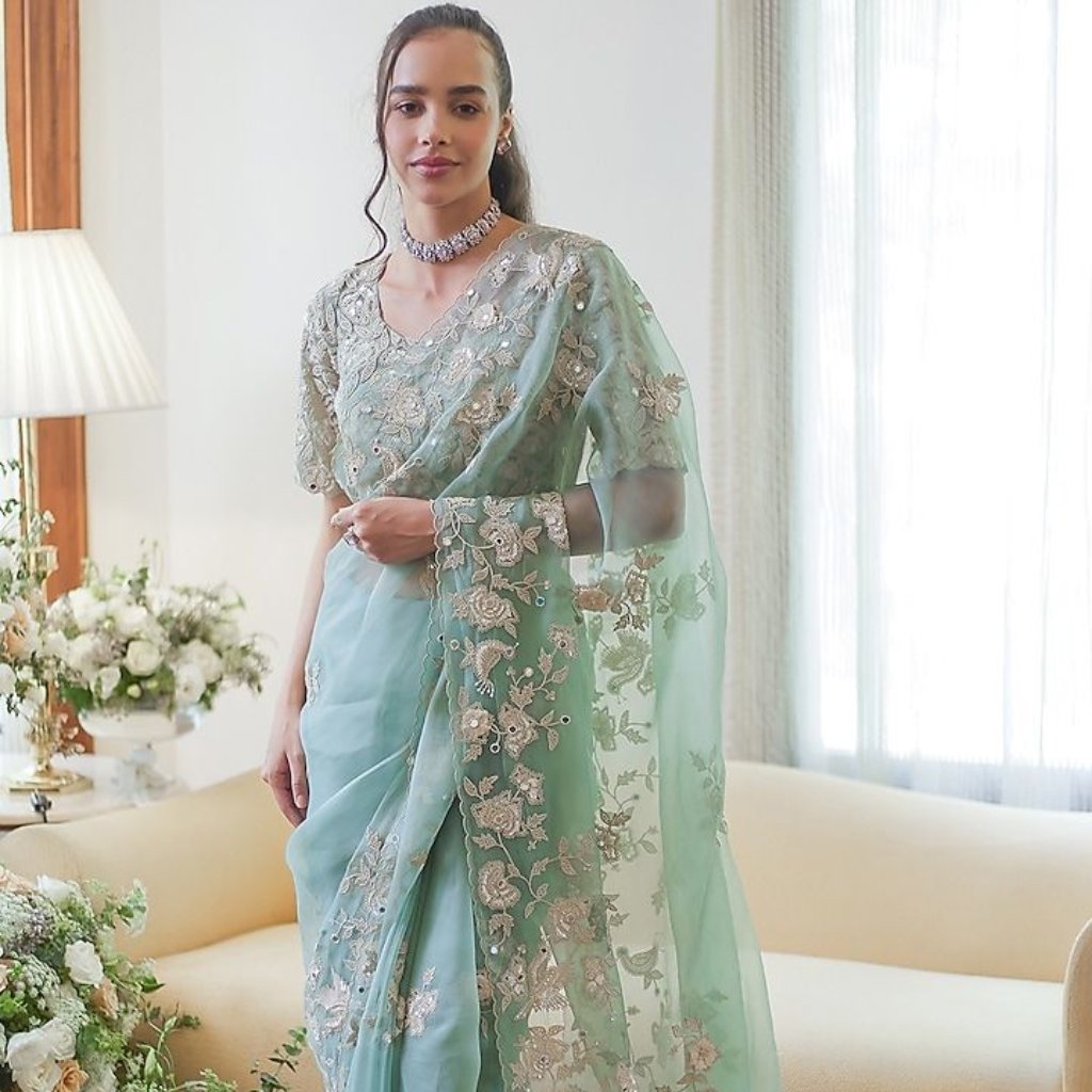 Green Organza Fabric: Elegant Choices for Fashion and Decor 18 Woman in a light blue-green saree with silver embroidery, standing indoors by a sofa and flowers.