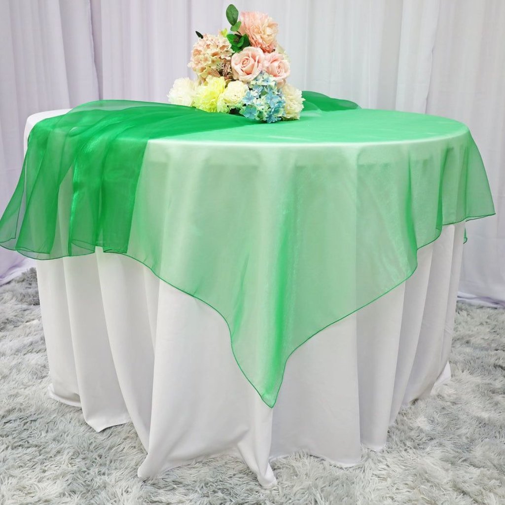 Green Organza Fabric: Elegant Choices for Fashion and Decor 20 A round table covered with a white tablecloth, topped with a bright green sheer overlay draped diagonally over the edges. A pastel floral arrangement sits at the center of the table. Light-colored drapes serve as a backdrop.