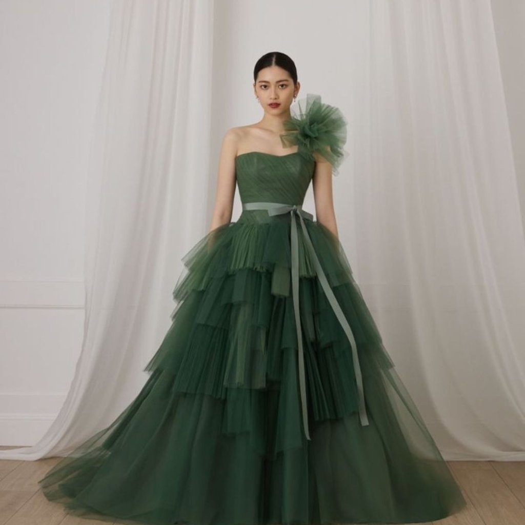 Green Organza Fabric: Elegant Choices for Fashion and Decor 16 A woman wearing an emerald green, one-shoulder ball gown with multiple layers of tulle, a sheer waist sash, and a large tulle flower on her shoulder. She stands indoors against a light curtain backdrop.