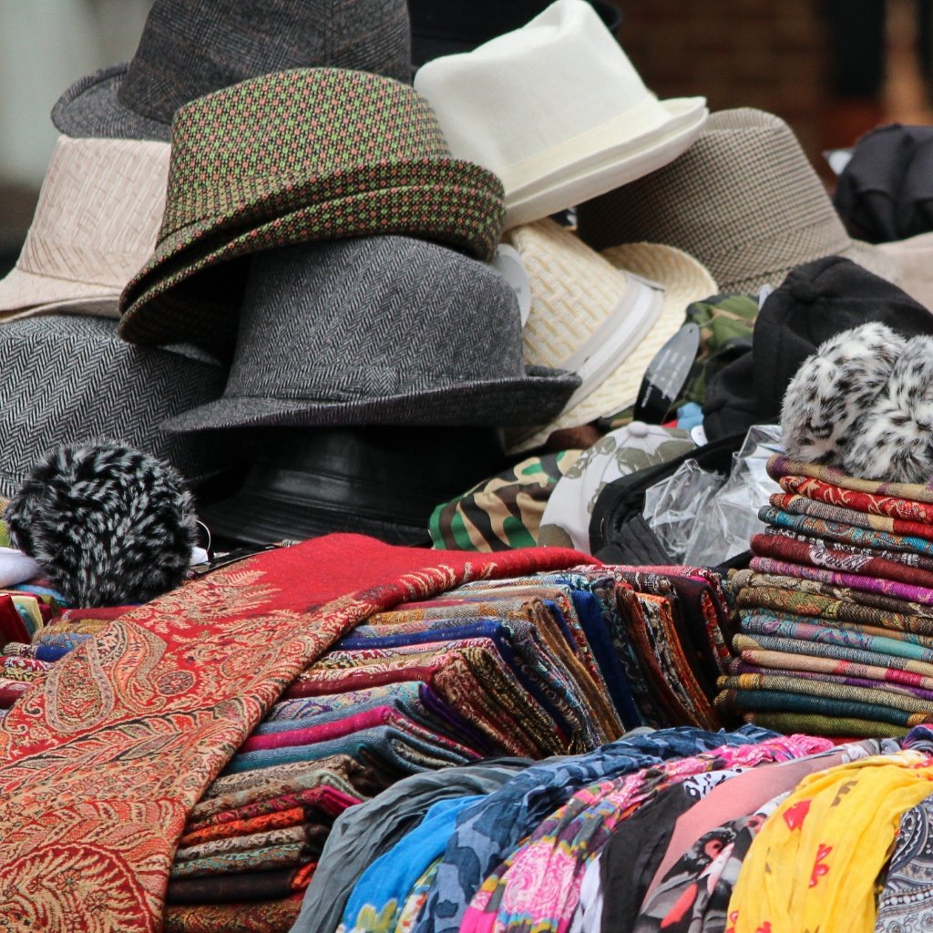 Why Seersucker Cotton Fabric Is Perfect for Summer Outfits 16 A colorful collection of hats and folded fabrics displayed on a market table, including felt fedoras, woven hats, and patterned scarves.