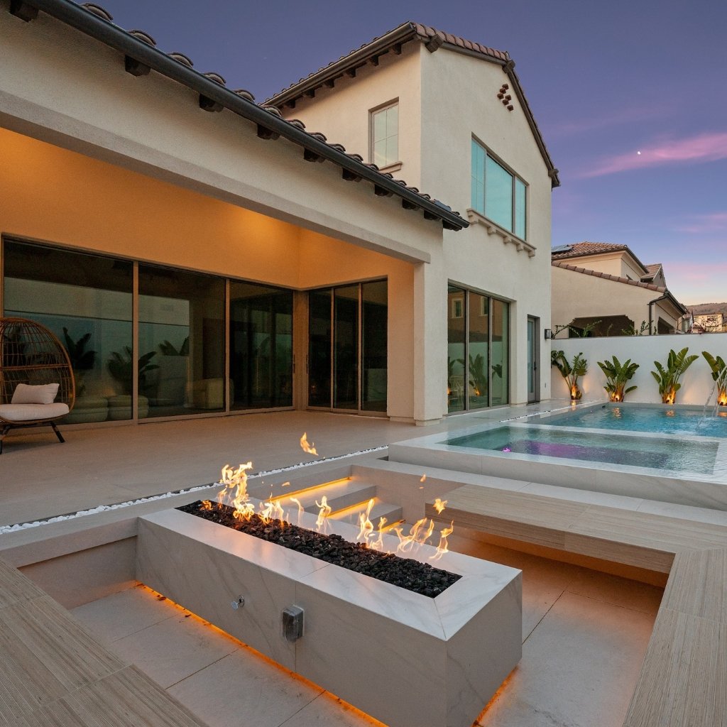 A modern backyard scene featuring a white, two-story house with large sliding glass doors, a rectangular pool, and an integrated rectangular fire feature with flames. The patio has light tan tiles, and there are small palm-like plants along a white perimeter wall. The sky is dusk-purple.