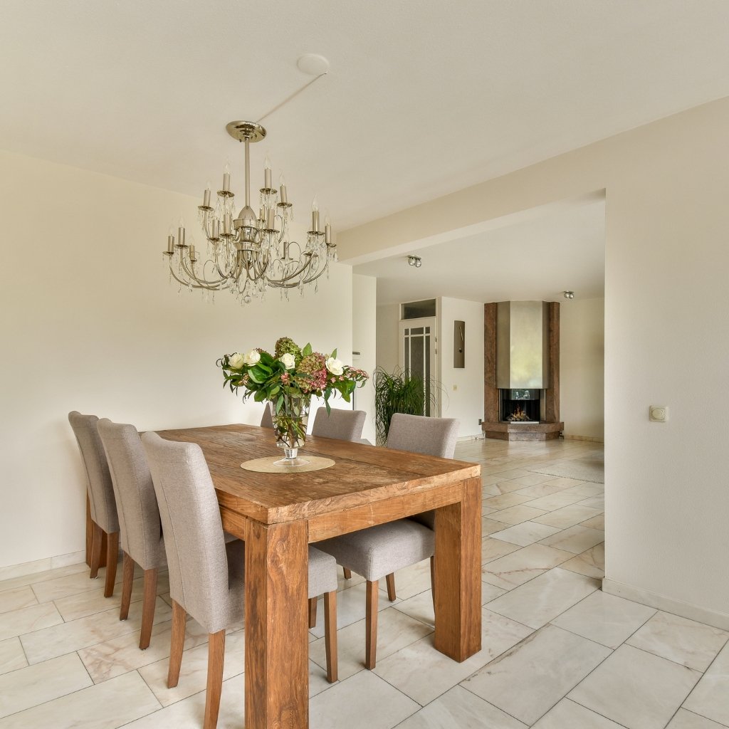 A bright dining room with a large wooden table and eight upholstered chairs. A crystal chandelier hangs above the table, and a vase of flowers sits in the center. The room has light-colored walls and tiled floors, with a modern fireplace visible in the adjacent living area.