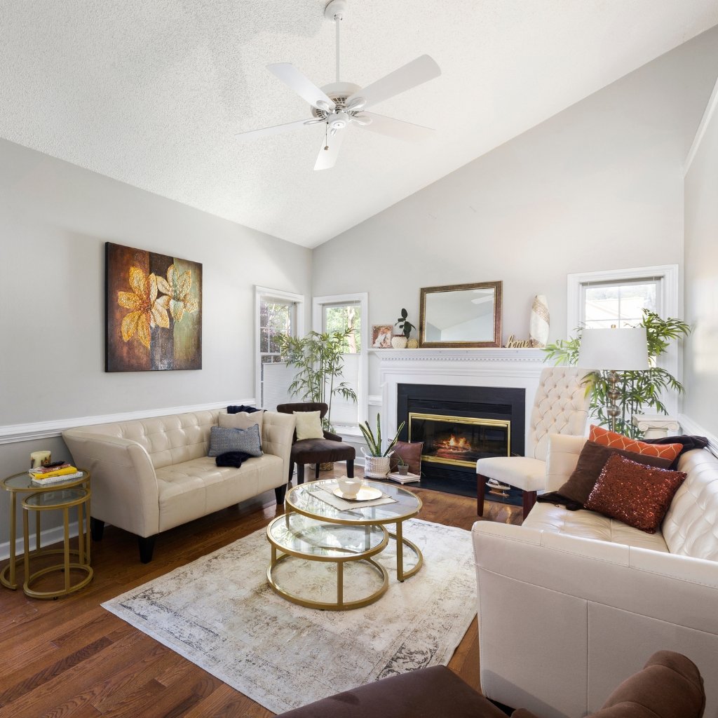 A bright, modern living room with light gray walls, a white ceiling fan, and a white fireplace. Cream tufted sofas face each other around a glass coffee table with gold frames. A cream rug sits on hardwood floors. Decorative pillows in brown and rust tones add color. Green potted plants flank the fireplace, and a large wall art piece with warm tones hangs on the left. Floor-to-ceiling windows and a mirror above the mantel reflect natural light.