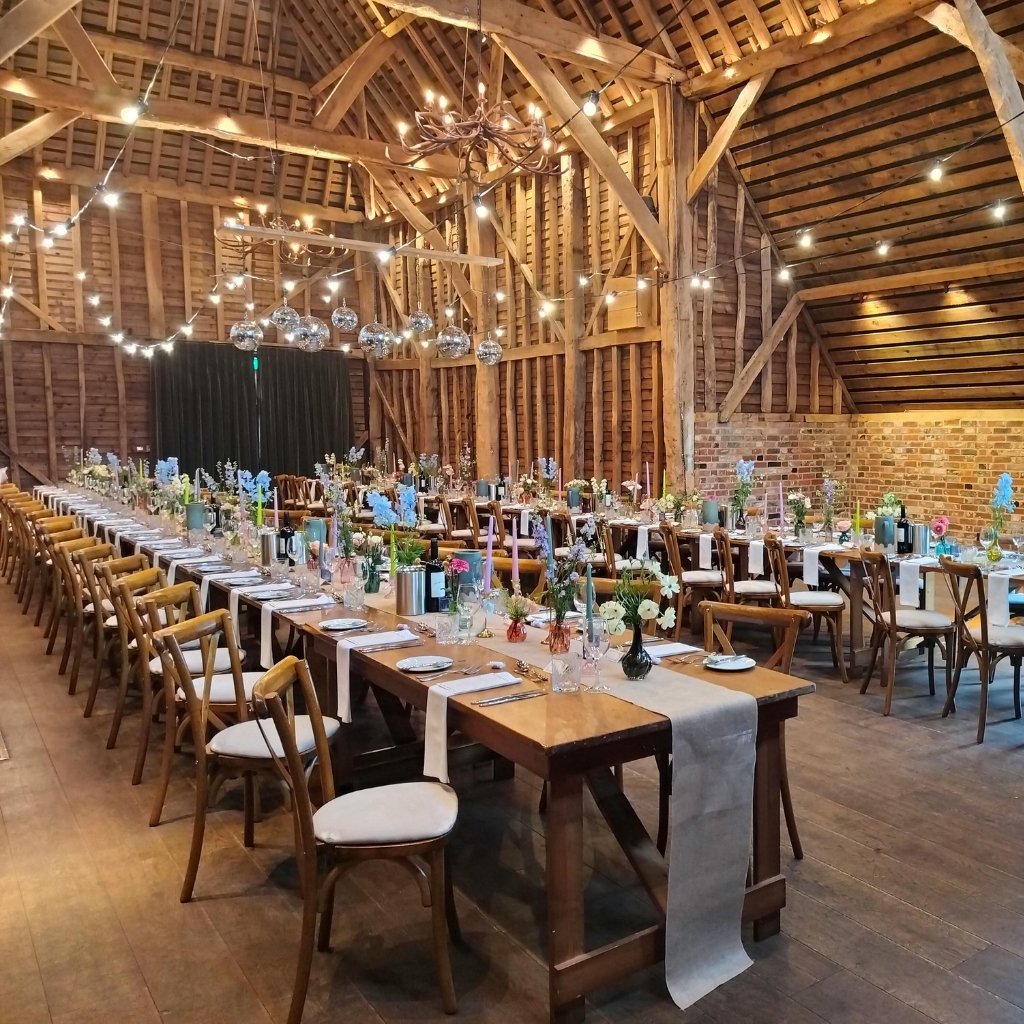 A rustic barn wedding setup with long wooden tables, neutral runners, and floral centerpieces. Warm string lights and disco balls hang from the ceiling, while wooden chairs line both sides of the tables.