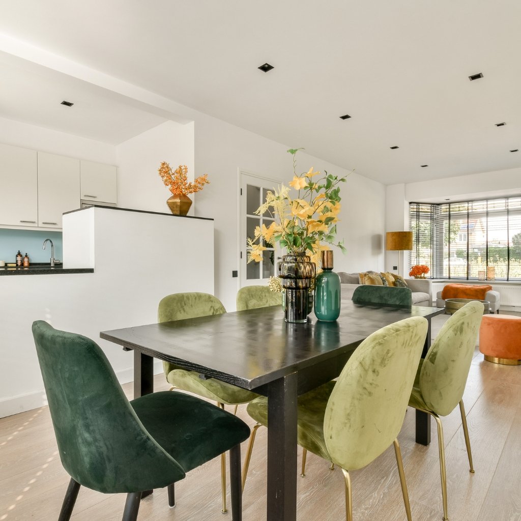 Interior dining room with a black wooden table surrounded by green and olive boucle wool chairs, centerpiece vases, and a bright, modern open-plan kitchen and living area.