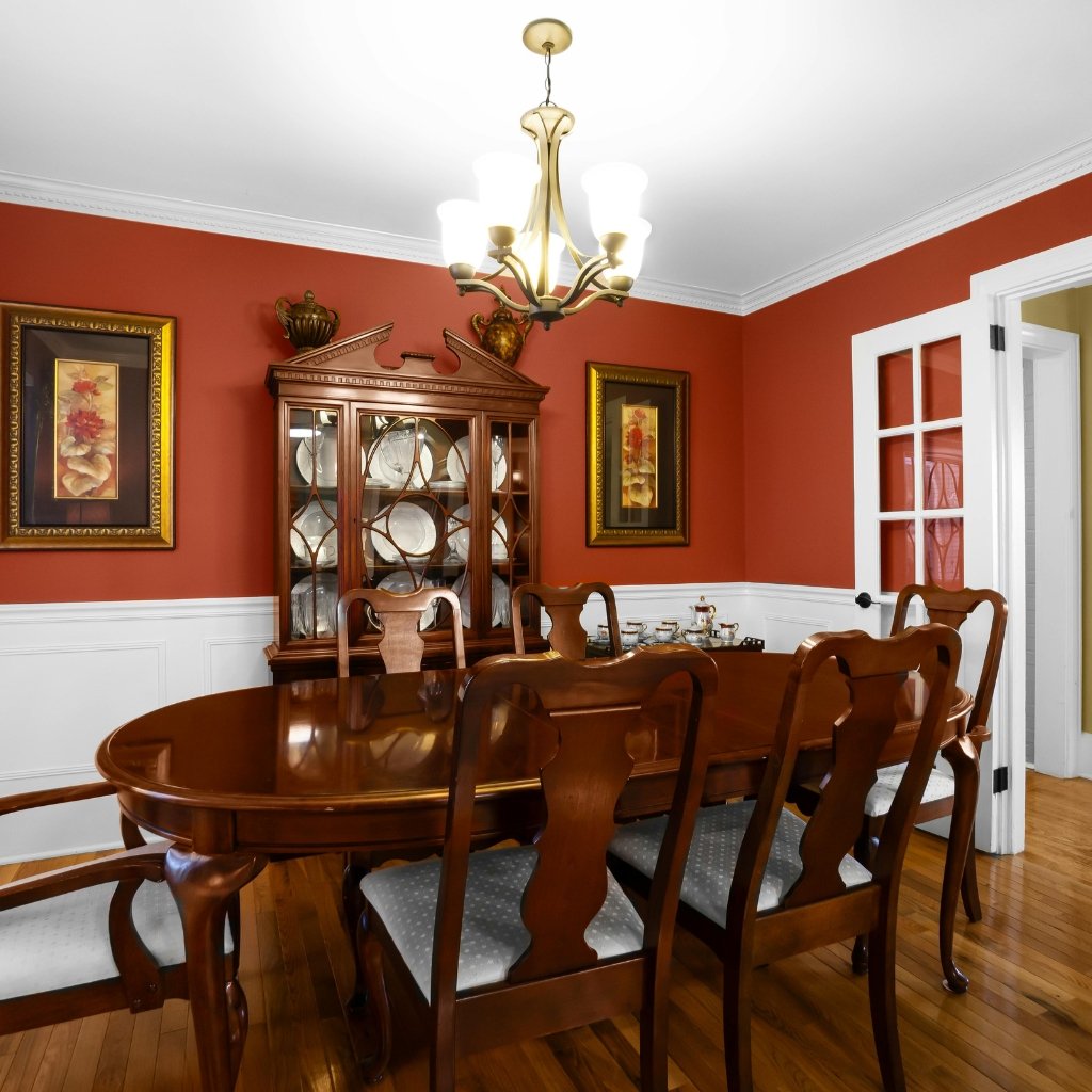 A traditional dining room with a polished wood table and chairs, red walls, a glass-front china cabinet, and a white wainscoted lower wall.