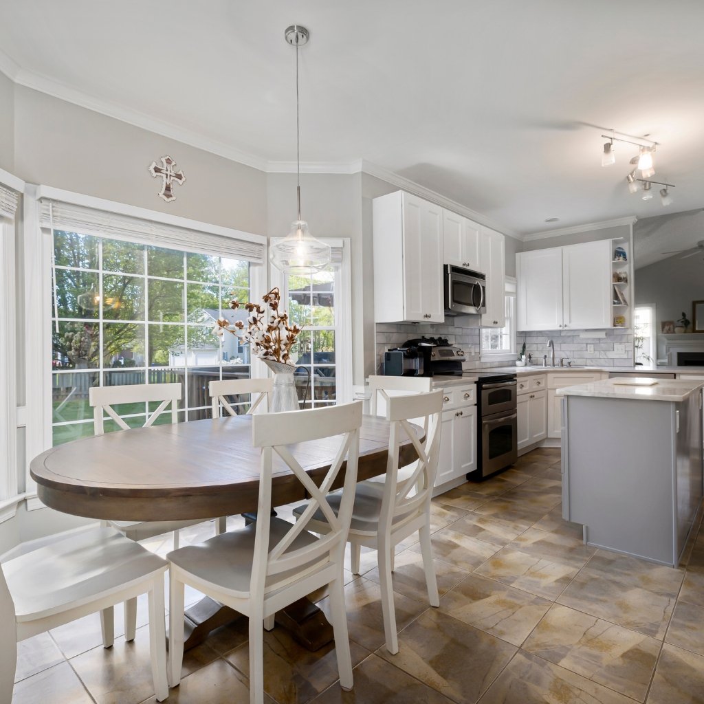 Bright kitchen and dining area with white cabinetry, a wooden round dining table, white chairs, large windows, and a modern stainless-steel stove; soft gray walls and tile floor.