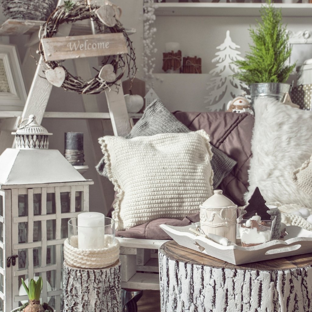 A cozy, neutral-toned living room decorated in a rustic-warm style with knitted, terry-wool textiles. A white knitted throw and plush cushions rest on a mauve sofa, with wooden stump tables and decorative seasonal accents around.