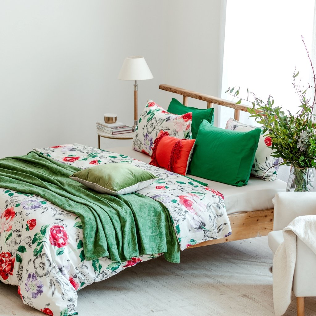 A cozy bedroom scene featuring a bed with a floral duvet, green and red accent pillows, and a green throw. A wooden headboard, a small side table with a lamp, and a vase of greenery sit nearby.