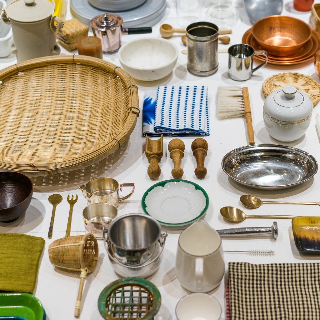 A diverse collection of kitchenware laid out on a white surface, including wicker baskets, wooden spoons, metal cups, ceramic bowls, brass utensils, cloth napkins, and various metal and ceramic dishes.