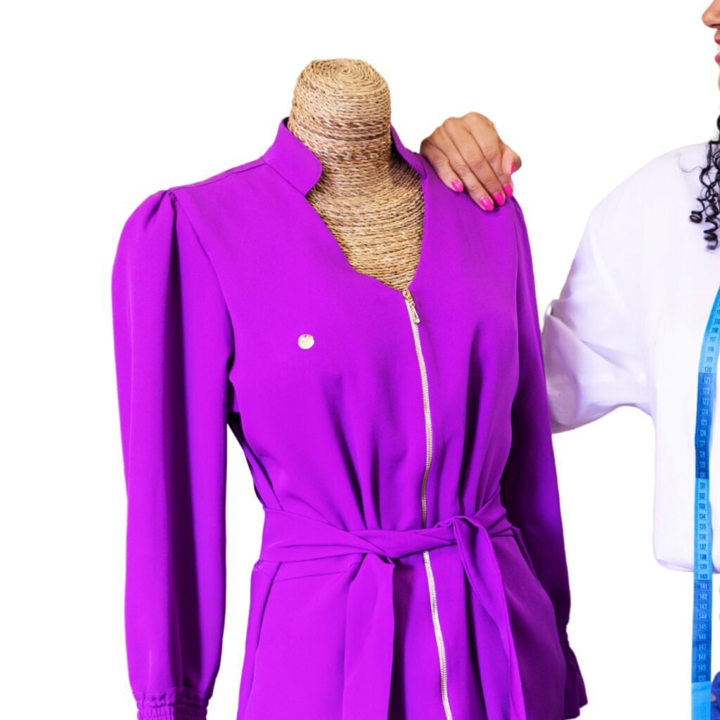 A bright purple knee-length belted blazer with a zip front on a mannequin. The garment has structured shoulders and long sleeves; a small gold pin is on the left chest. The mannequin is standing next to a person holding a measuring tape.
