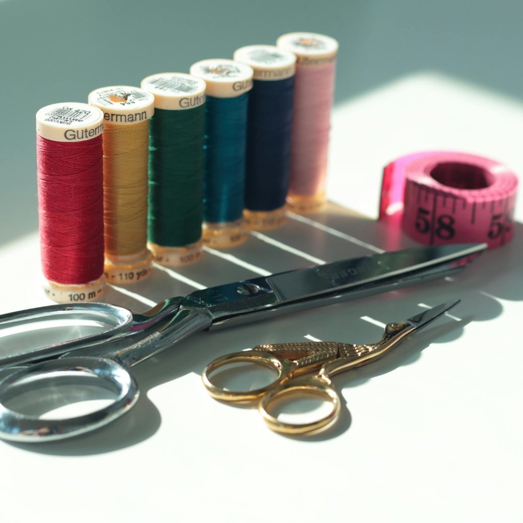 A row of colorful spools of thread (red, yellow, green, teal, blue, pink) standing upright on a white surface, with a pair of silver scissors, gold-tone thread-cutting snips, and a pink measuring tape in the foreground. Bright, natural light casts soft shadows.
