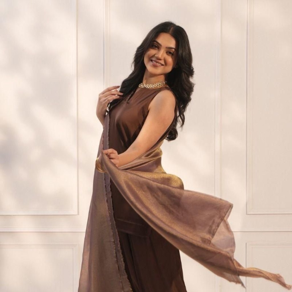 A woman wearing a brown sleeveless dress with a shimmering brown-to-golden scarf, posing gracefully in a well-lit room with white paneled walls.
