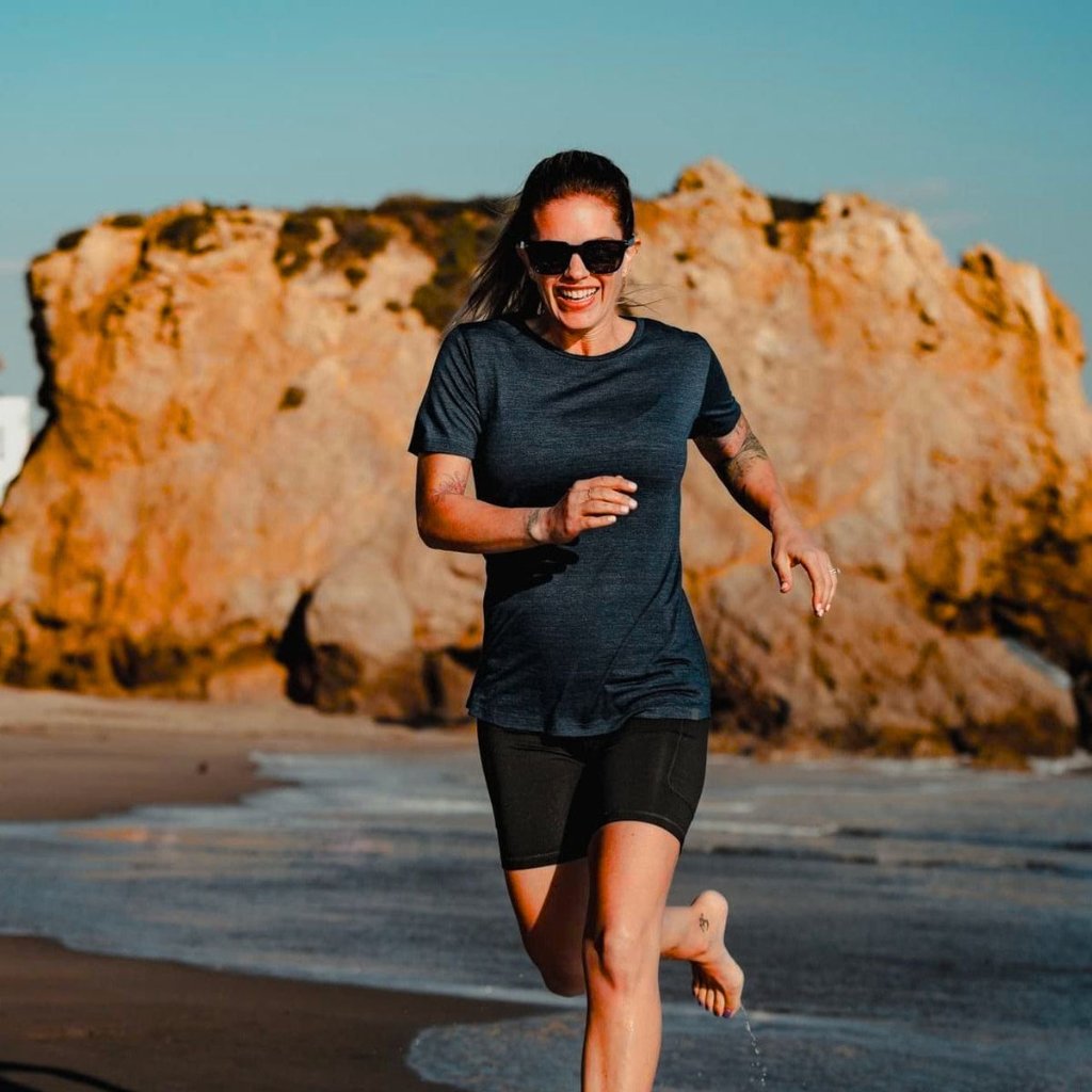 A woman wearing a dark gray T-shirt and black shorts is running along a beach at the edge of rocky cliffs. She’s smiling, wearing sunglasses, and the sun casts warm light on the scene.