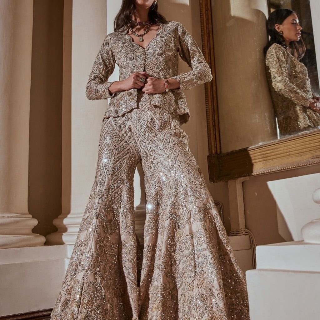 A woman wearing an ornate, champagne-beige lehenga with heavy sequin and bead embroidery. The outfit features a long-sleeved, fitted jacket over a full-length, flared skirt. She stands beside a large, framed mirror in a classical-colonnade setting, accessorized with layered necklaces and bracelets.