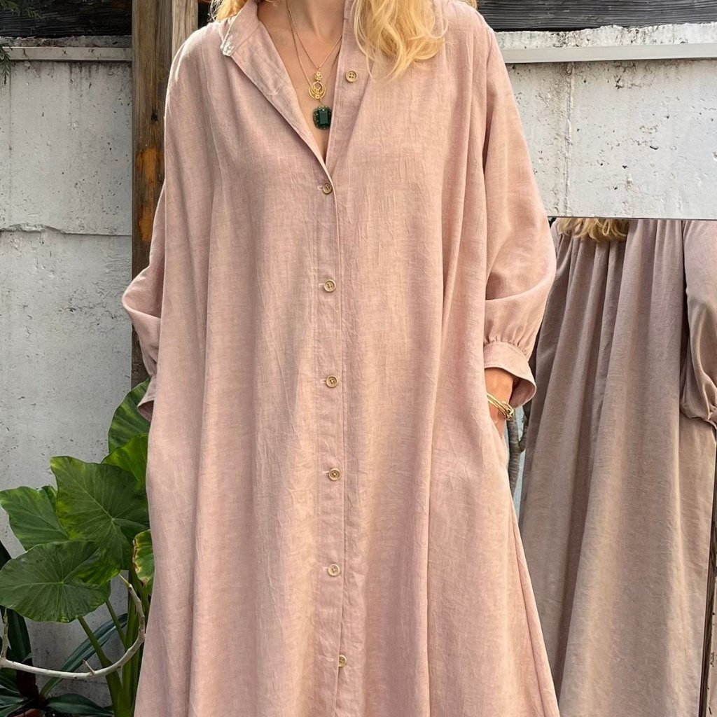 A knee-length, loose-fitting button-down dress in a soft dusty pink linen. The garment features a relaxed collar, long voluminous sleeves gathered at the cuffs, and light wooden buttons running down the front. A woman’s torso and part of her legs are visible; she wears layered gold necklaces and bracelets, and her hand rests in a side pocket. The background shows a weathered wall and a mirror reflecting the dress.