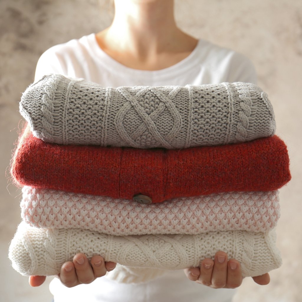 A person in a white top holds a stack of knitted sweaters in gray, red, pink, and white.