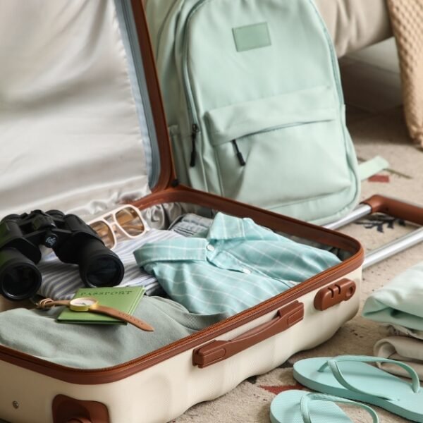A vintage-style open suitcase on a carpet, with neatly packed clothes and accessories. Inside the case are a green neck pillow, a pair of binoculars, glasses, and small travel items. A mint-green backpack sits nearby and folded clothes and flip-flops are arranged on the floor. Merino Travel Clothes