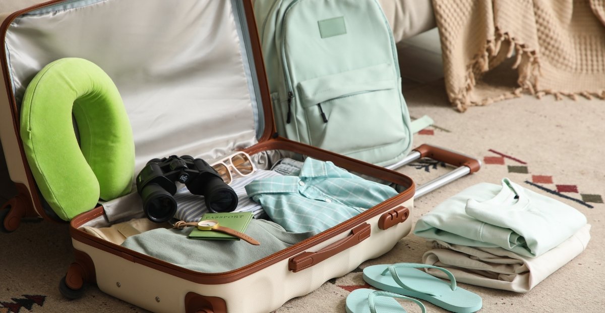 A vintage-style open suitcase on a carpet, with neatly packed clothes and accessories. Inside the case are a green neck pillow, a pair of binoculars, glasses, and small travel items. A mint-green backpack sits nearby and folded clothes and flip-flops are arranged on the floor. Merino Travel Clothes