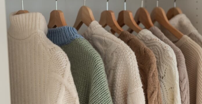 A row of knit sweaters hanging on wooden hangers in a closet, featuring cream, light green, blue, and beige tones. Merino Wool Clothing
