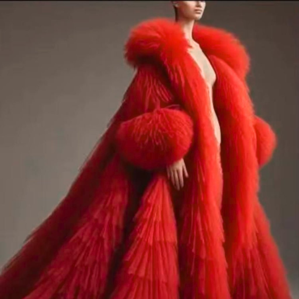 A fashion model wears a dramatic red fur coat with oversized puffs and long, flowing layers; the coat envelops most of her body and the model stands against a neutral backdrop.