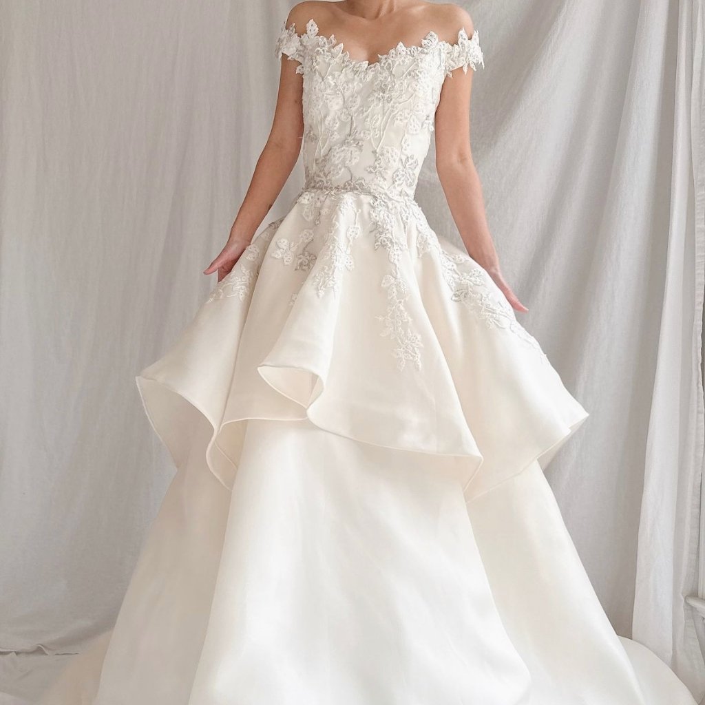 A bridal gown in ivory organza with an off-the-shoulder neckline and delicate floral lace appliqués. The dress features a fitted bodice and a voluminous, multi-tiered skirt with sculpted, cascading ruffles that create movement and drama.
