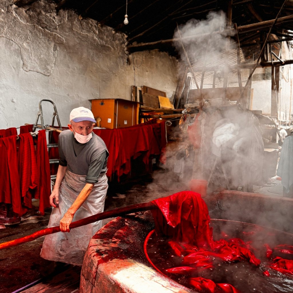 A man wearing a cap, face mask, and apron stands near a large vat of red fabric or liquid, stirring with a long wooden paddle in a workshop or factory setting. Steam rises from the vat, and red garments hang along a rack in the background.