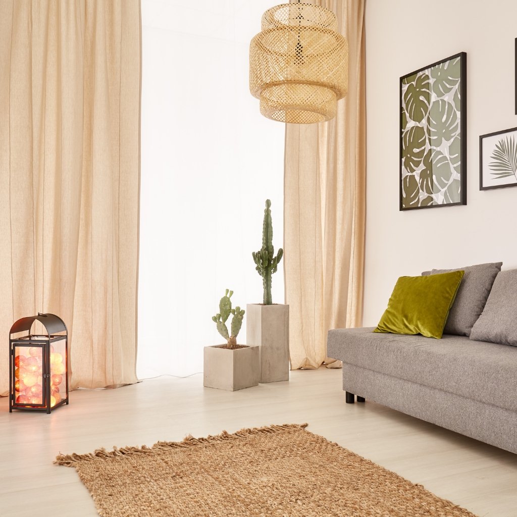 A bright, cozy living room with beige curtains, a woven hanging light, and a gray sofa with a lime-green cushion. Two potted cacti in concrete blocks sit on the floor near the window, while a woven rug lies on light wood flooring. A lantern with warm light sits to the left, and framed botanical prints adorn the wall.
