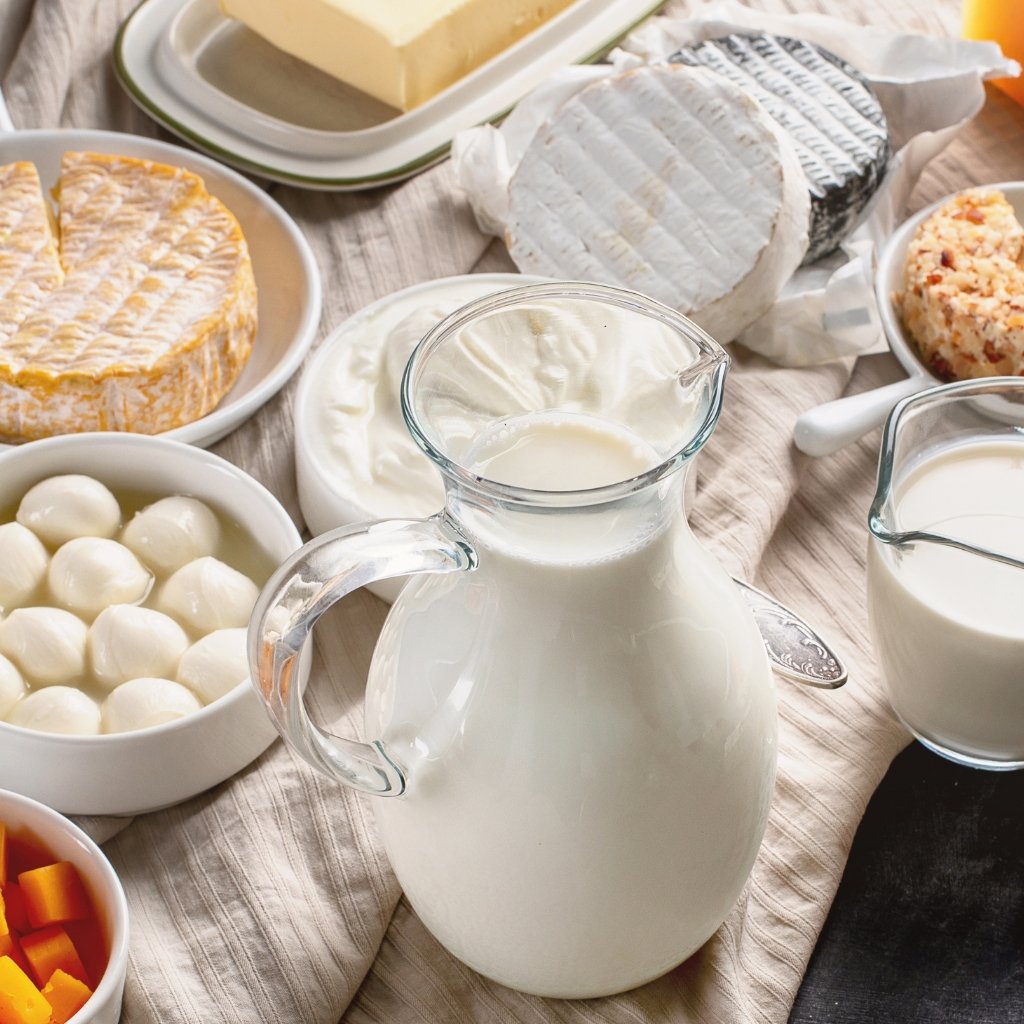 A still-life dairy spread on a light striped cloth, including a glass pitcher of milk, a small pitcher of milk, a bowl of white cheese balls, various cheeses on plates, a wheel of soft cheese wrapped in white rind, and small bowls with cut vegetables. The scene sits on a wooden surface.