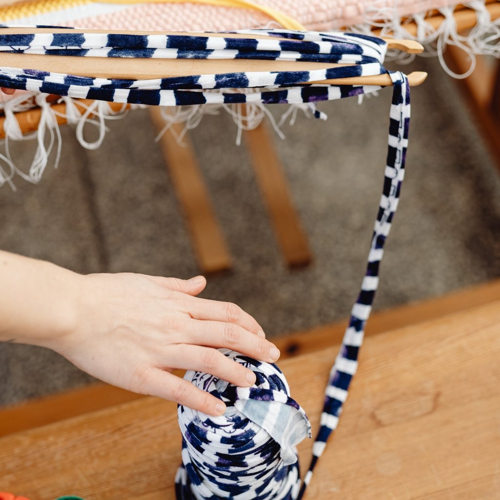 A close-up of a hand winding a striped blue-and-white cord on a wooden loom. The fabric strands and fringe loom overhead, with the hand grasping a rolled section of the woven material.